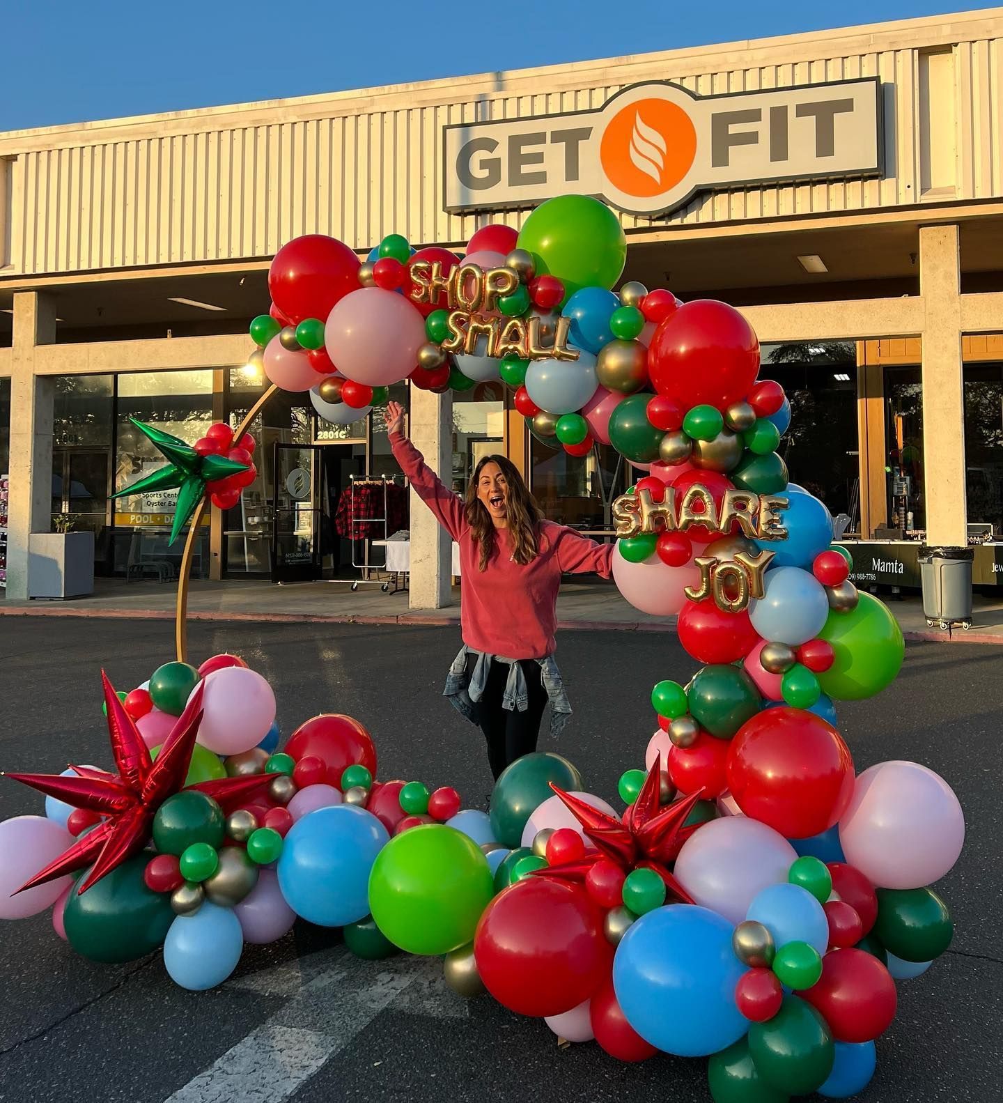 Woman with arms raised poses in front of a balloon arch with the words 