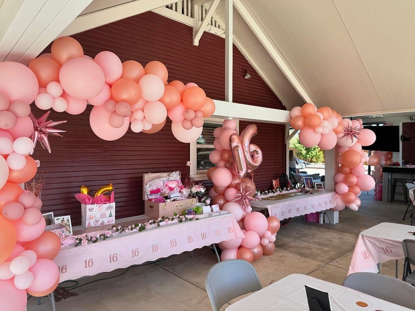 Pink balloon arch decorates a sweet sixteen party at an outdoor pavilion. Tables with gifts and decor.