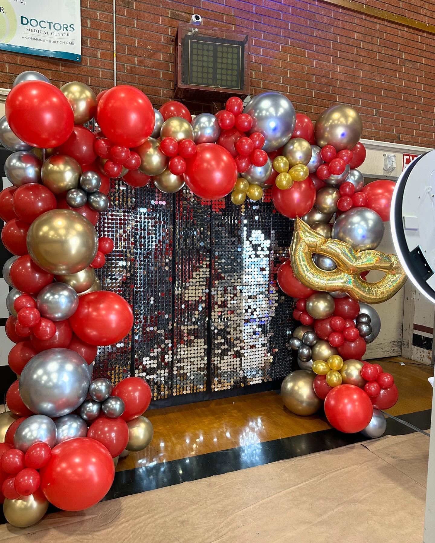 Red, gold, and silver balloon arch frames a mirrored wall, with a gold mask balloon.