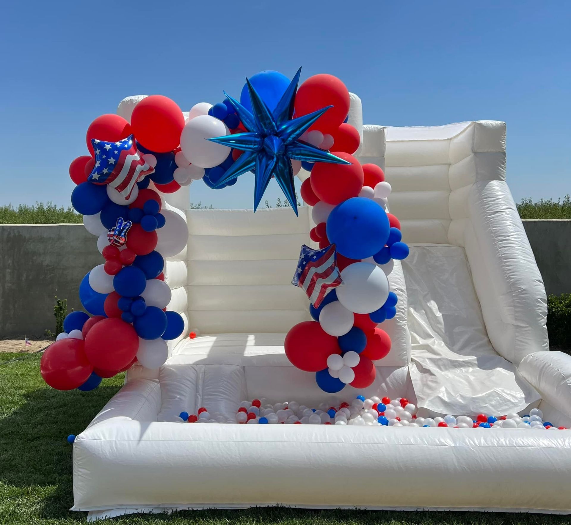 White inflatable slide with red, white, and blue balloon arch in an outdoor setting with a blue sky.