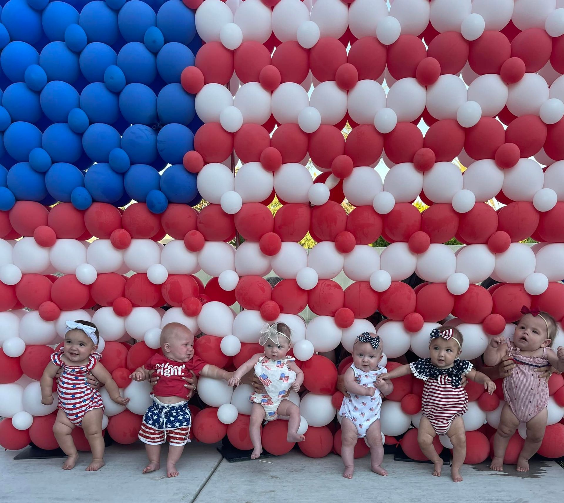 Seven babies dressed in patriotic outfits stand in front of a balloon American flag.