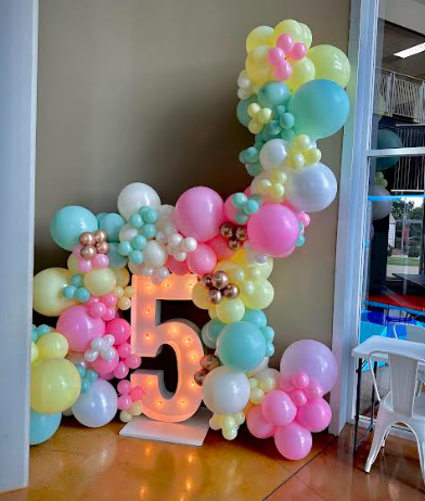 Balloon arch with a lit-up number 5. Pastel colors: pink, yellow, blue, and white.