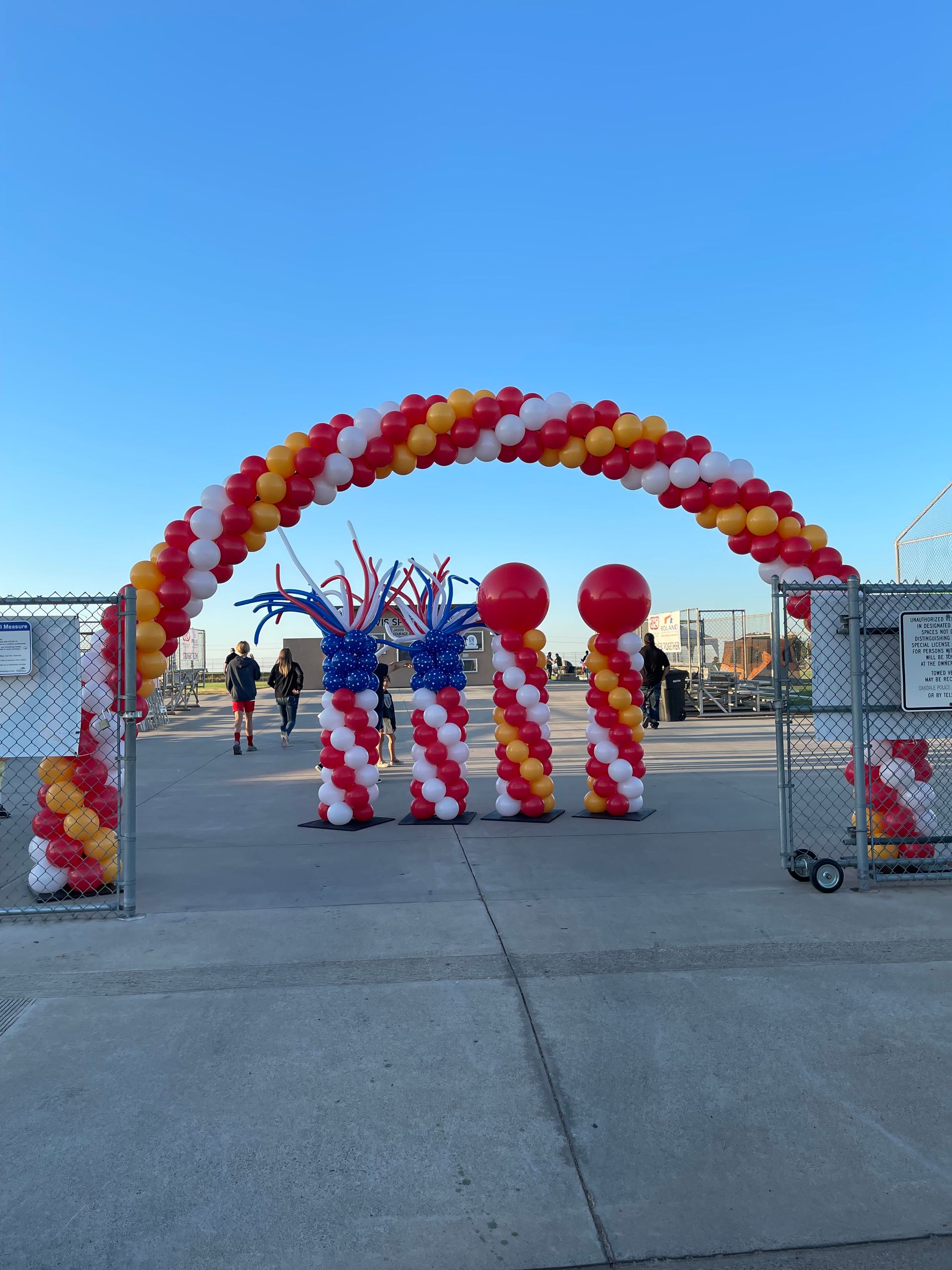 Balloon arch entrance with red, yellow, and white decorations. People are walking on the concrete.