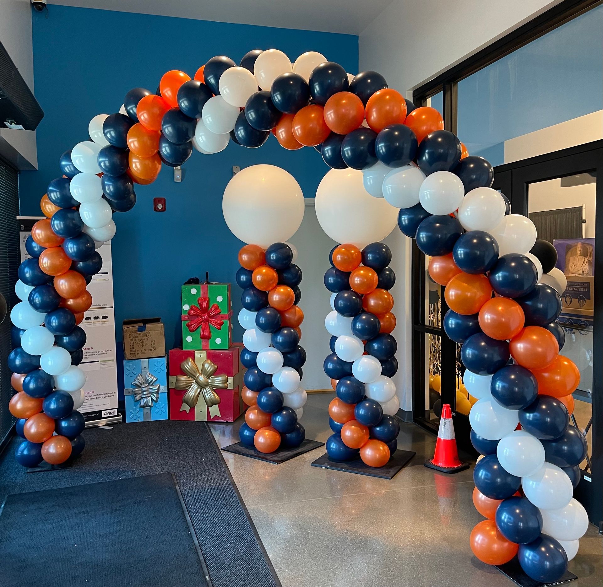 A festive balloon arch and pillars in orange, white, and navy, welcoming guests into a building.