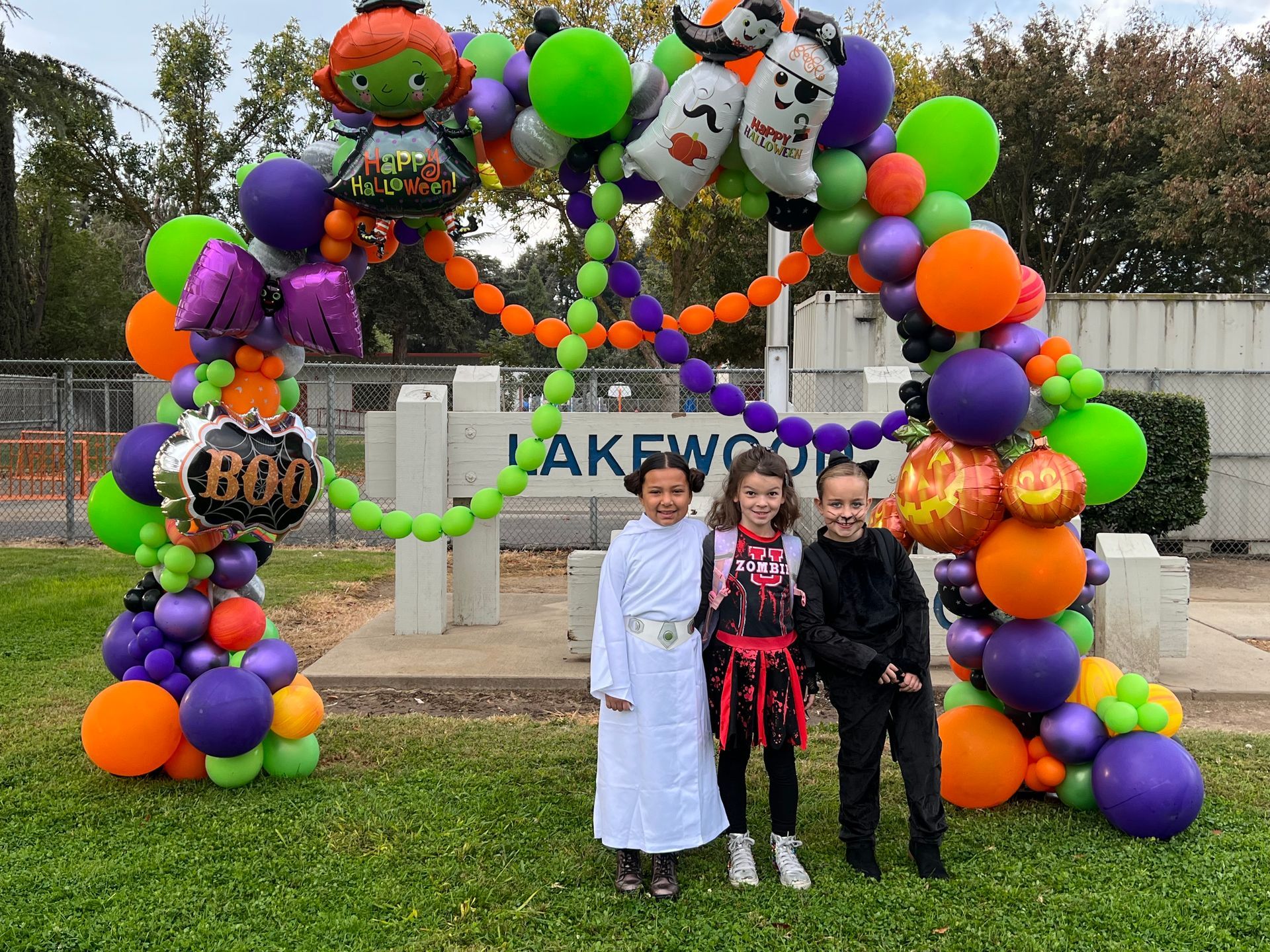Three kids in Halloween costumes pose under a balloon arch in front of a school sign.
