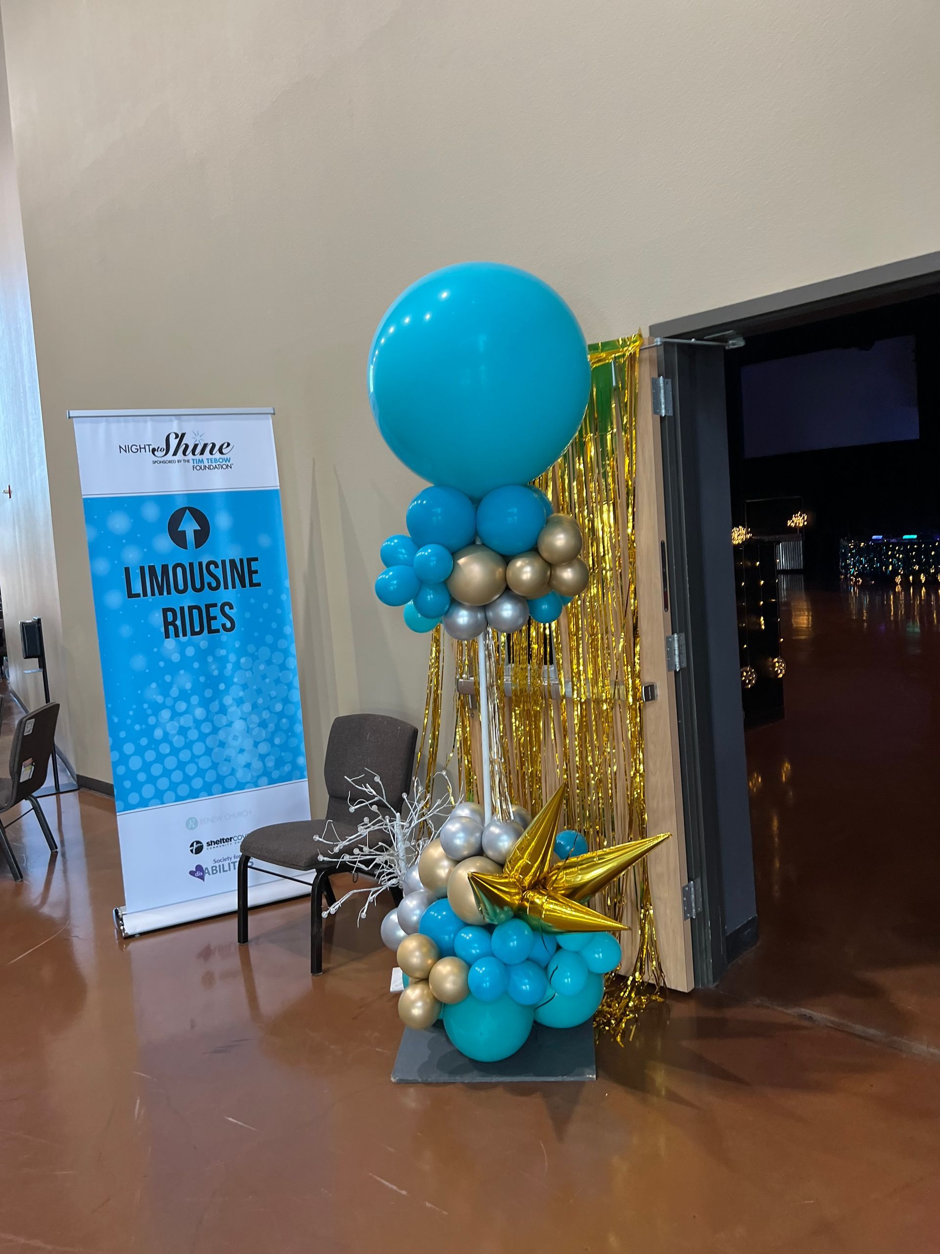 Balloon arch with teal and gold balloons, near a banner, chair, and doorway.