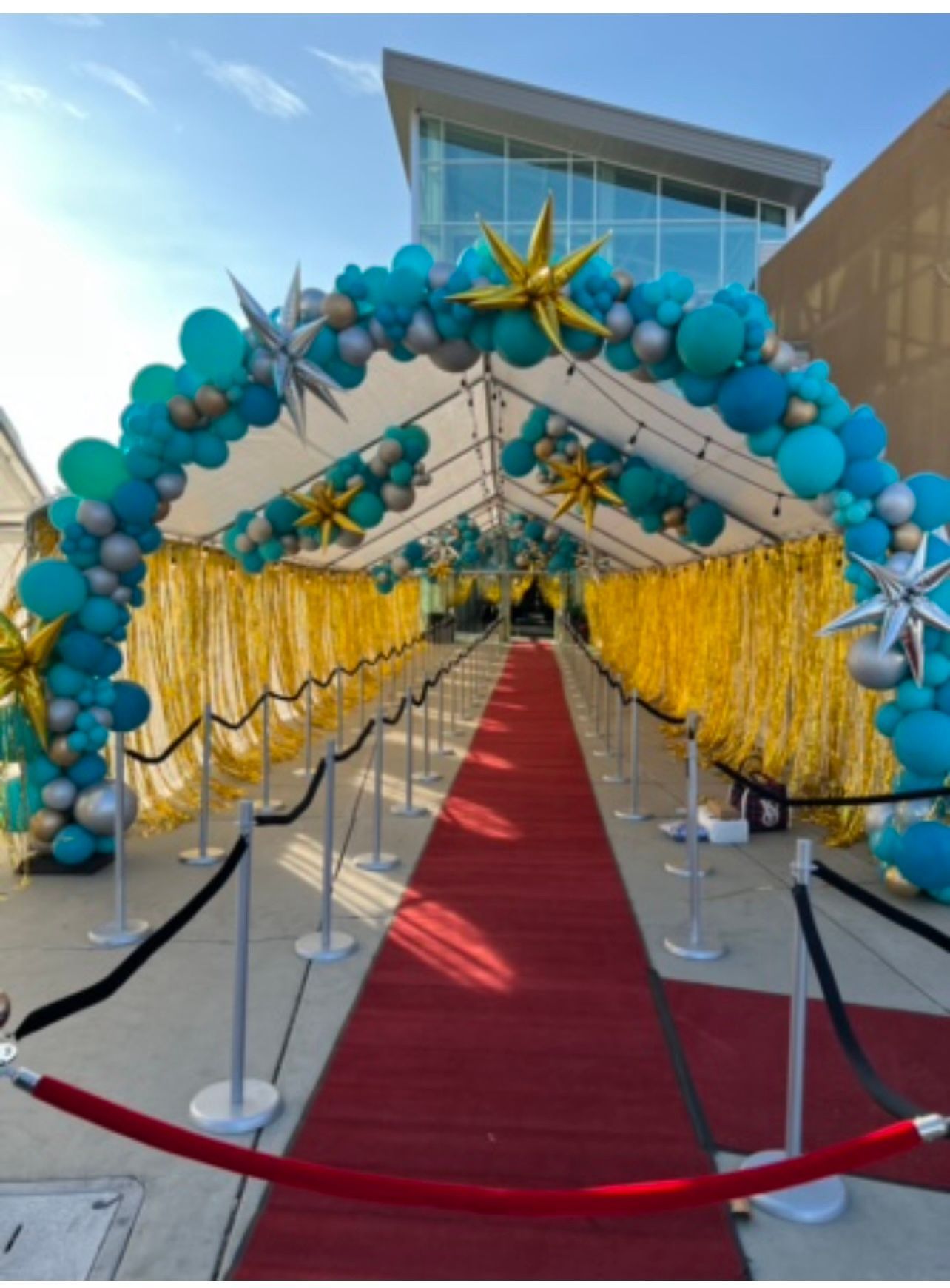 Red carpet entrance, turquoise and silver balloon arch, gold star decorations, gold fringe backdrop.