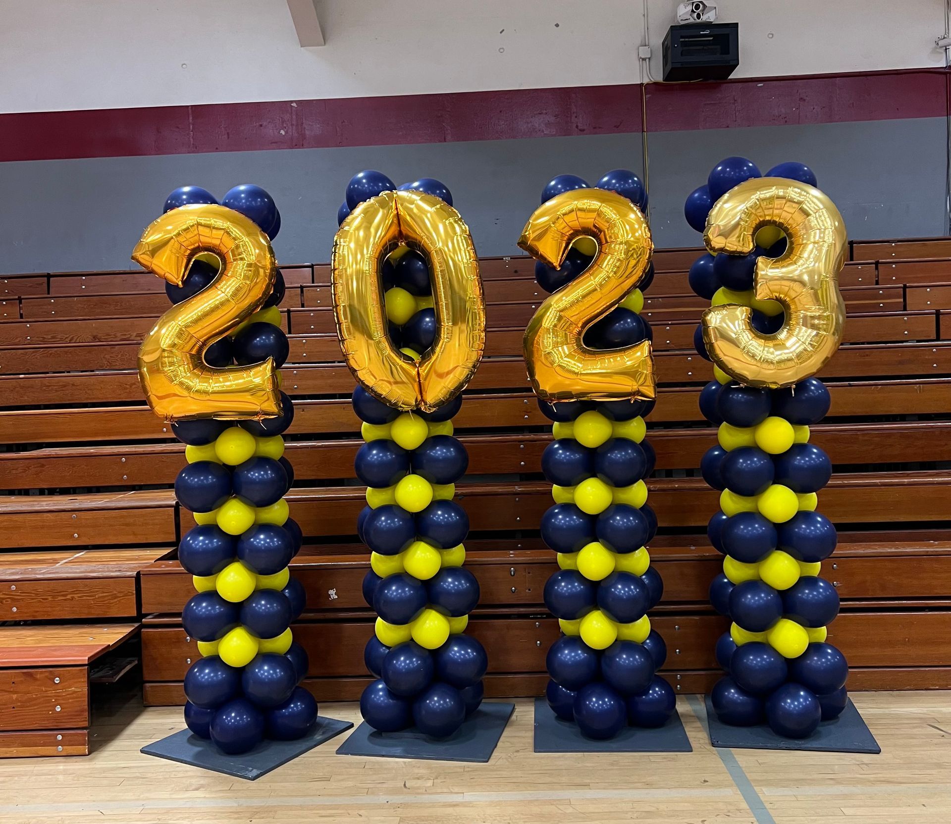 Graduation year 2023 made of gold balloons atop blue and yellow balloon columns, set in a gymnasium.
