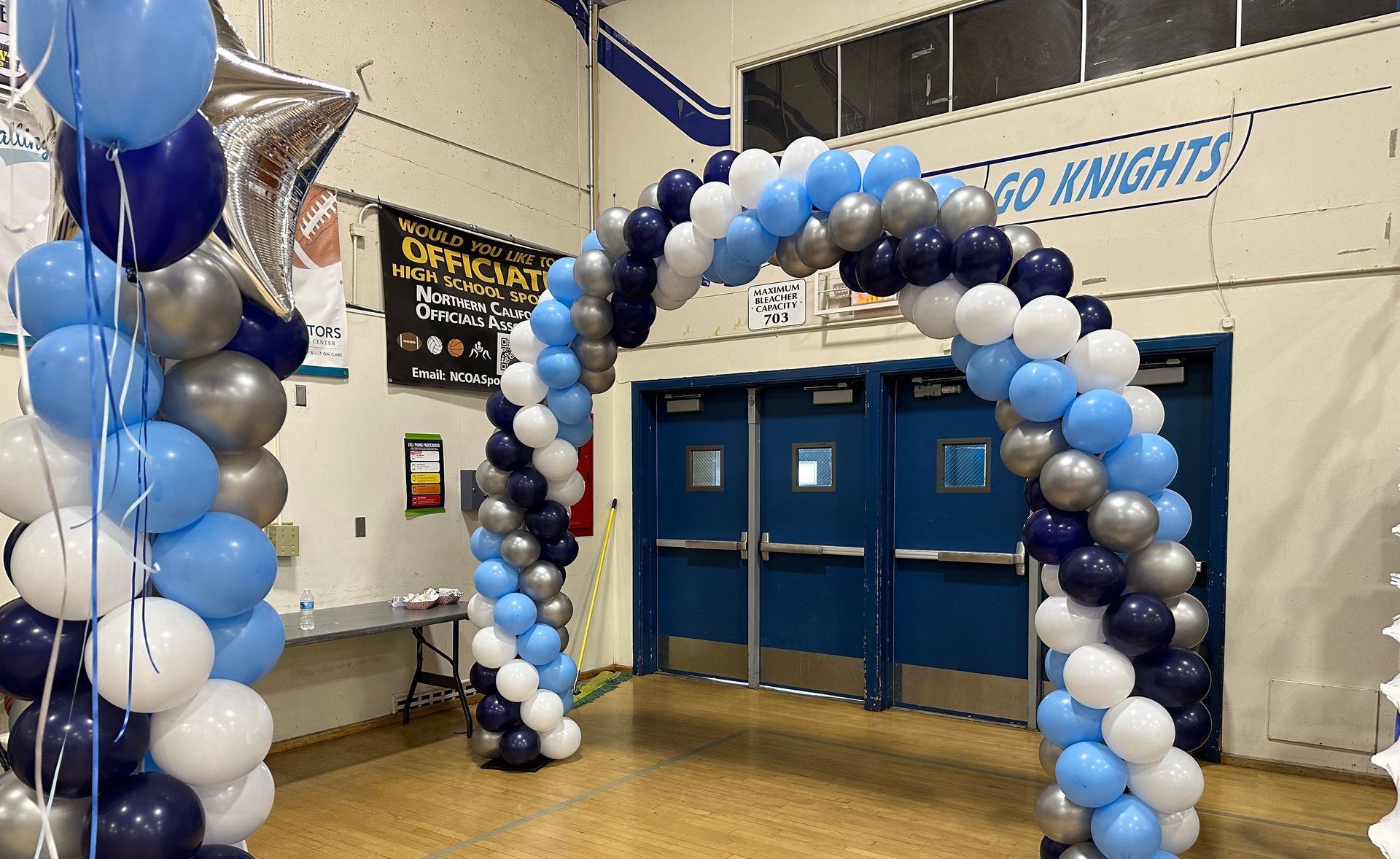Balloon arch and pillar in school gym; blue, silver, and white balloons decorate entrance.