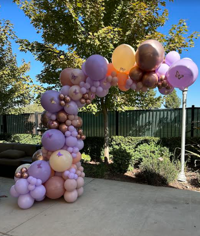 Balloon arch in shades of pink, purple, and gold, set outside.