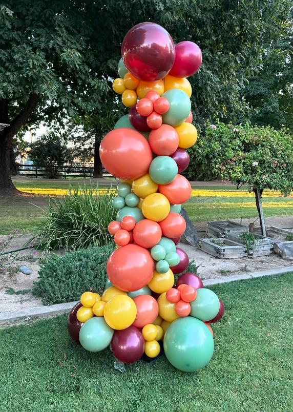 Balloon arch in autumn colors: orange, yellow, green, and burgundy, on grass, outdoors.
