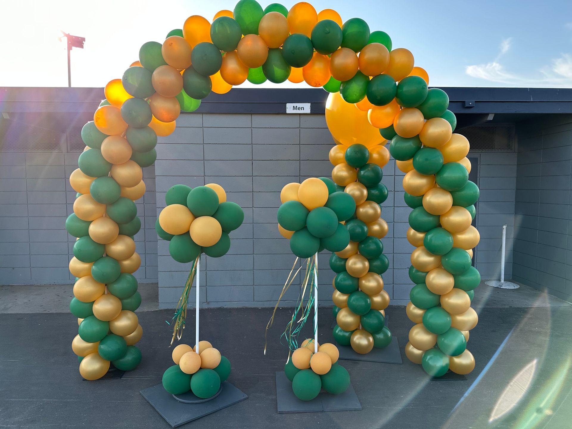 Balloon arch and topiary decorations in green and gold at an outdoor event.