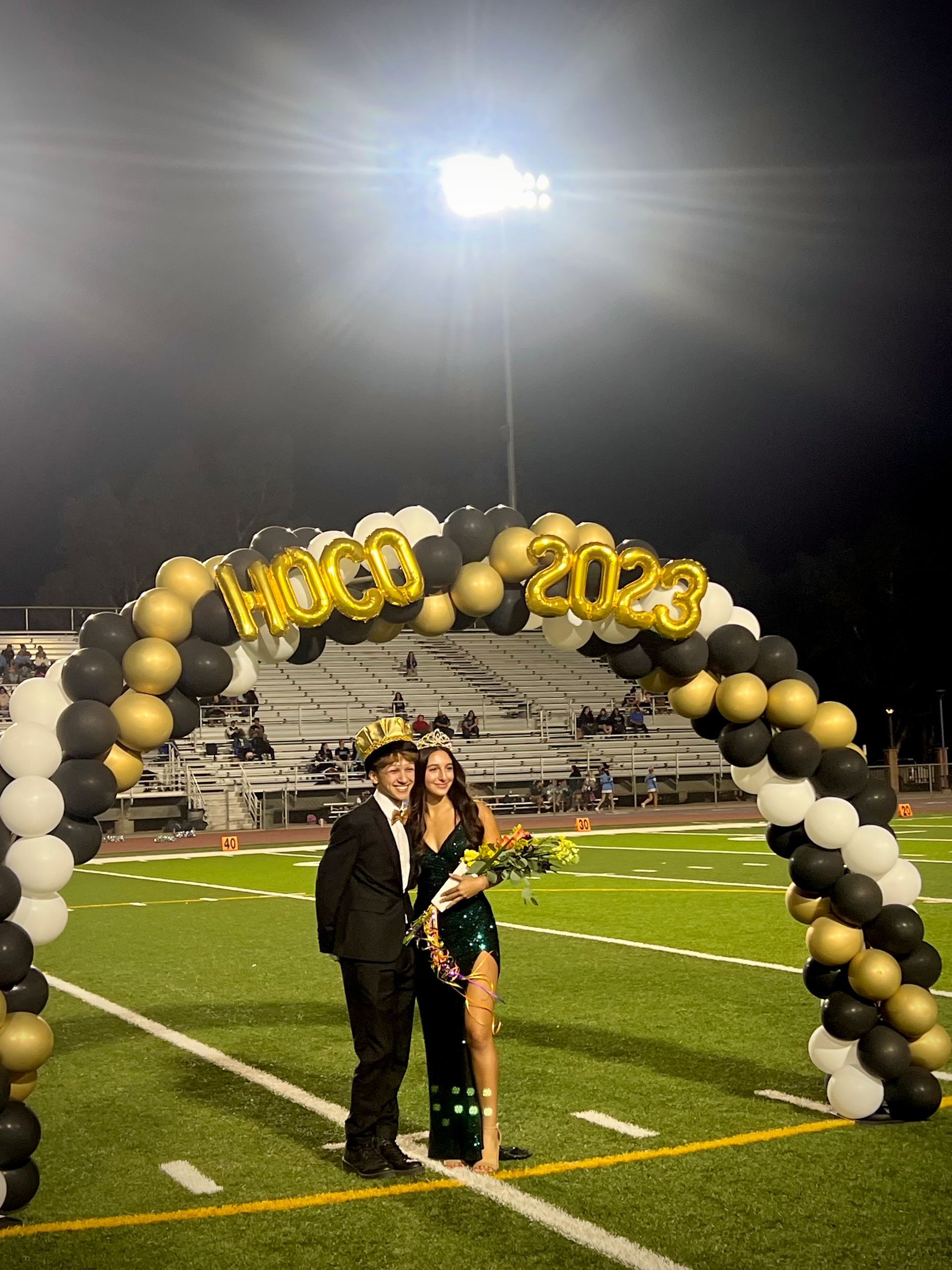 Homecoming king and queen under balloon arch, smiling, on a football field at night.