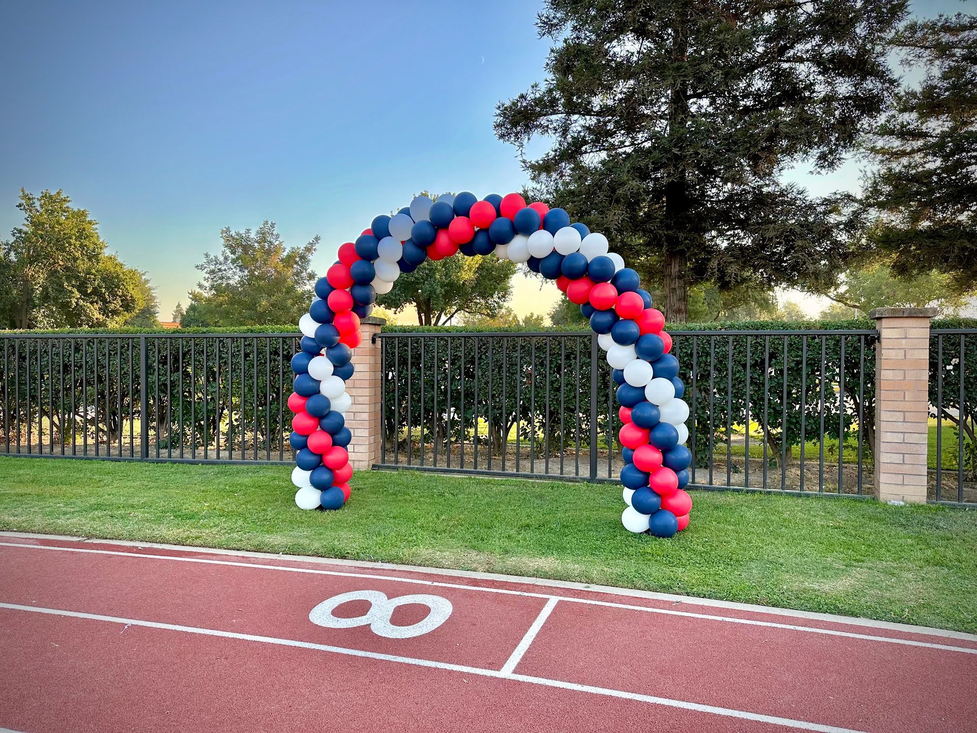 Balloon arch in red, white, and blue over a track, near a fence and trees.