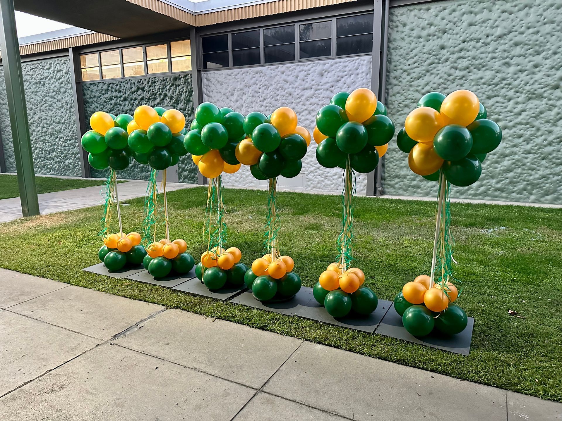 Green and gold balloon trees line a sidewalk, set on black bases, outside a building with a stone wall.