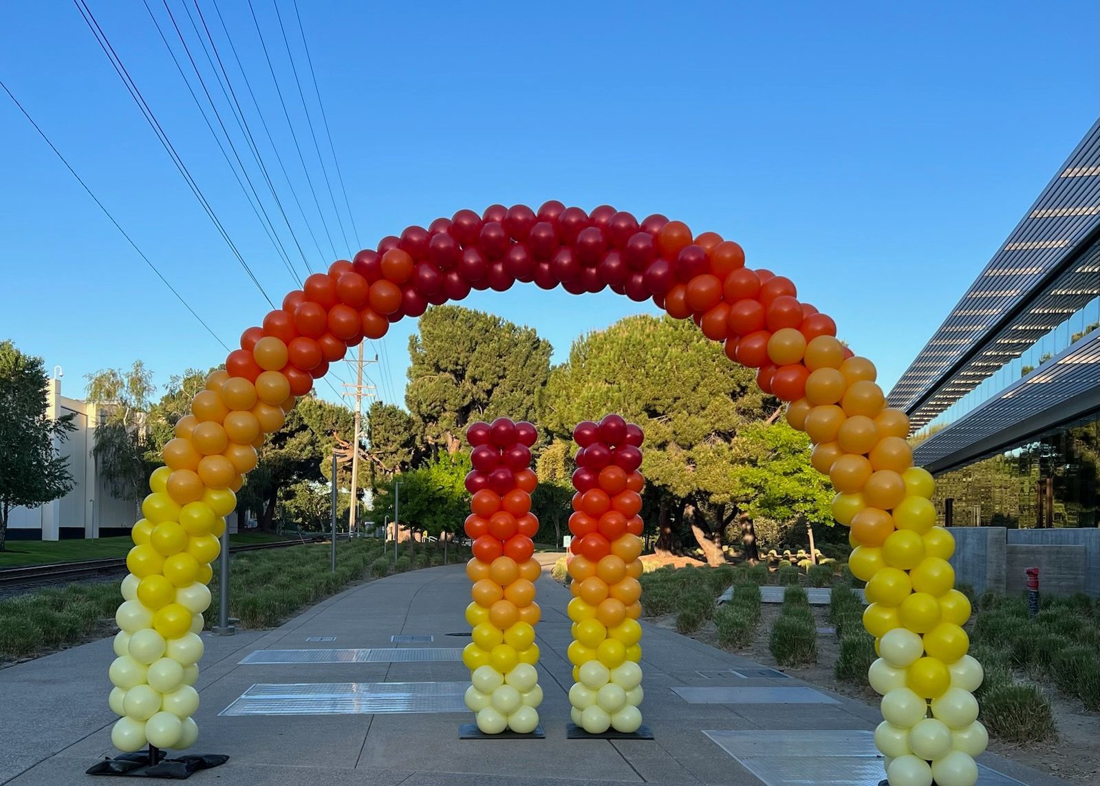 Balloon arch and columns in a gradient of yellow to red, set outside with trees and a modern building.