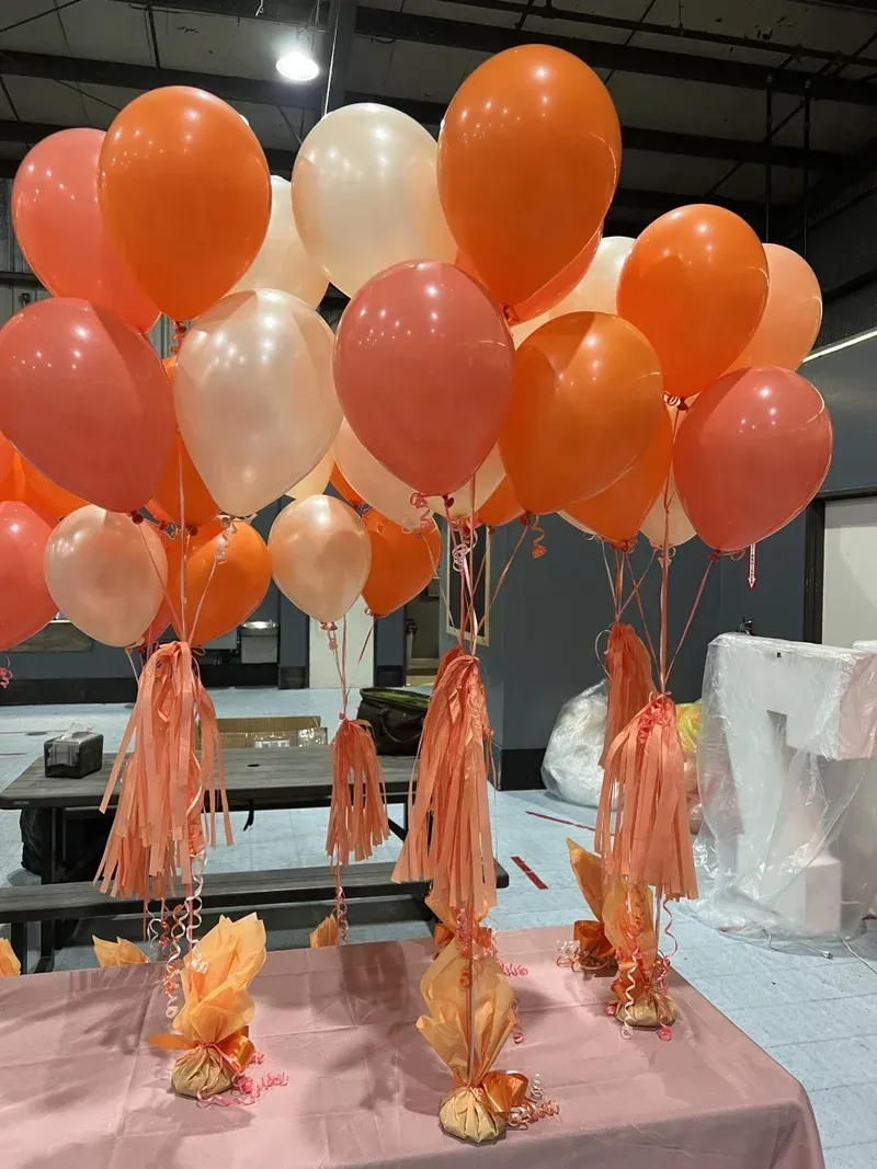 Balloons in orange, peach, and white with tassels on a pink tablecloth, likely for a celebration.