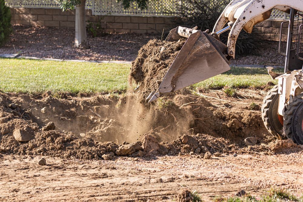 Skid steer digging in a yard, dumping dirt onto a pile in the sunlight.