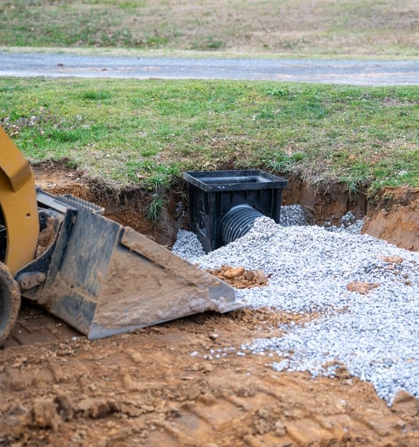 A black drainage structure and pipe surrounded by gravel, next to a backhoe in an open trench.