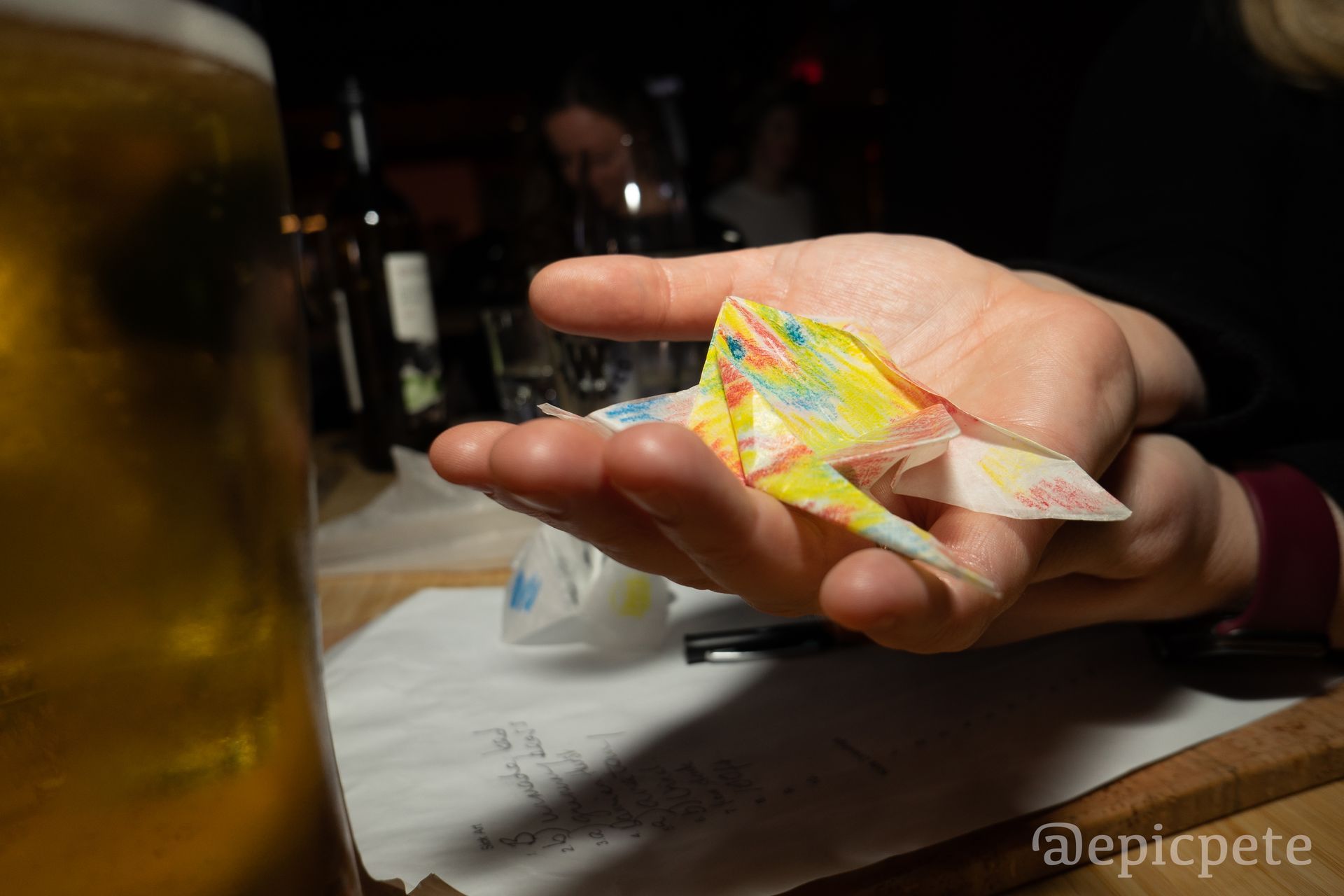 A person is holding an origami bird in their hand next to a glass of beer.