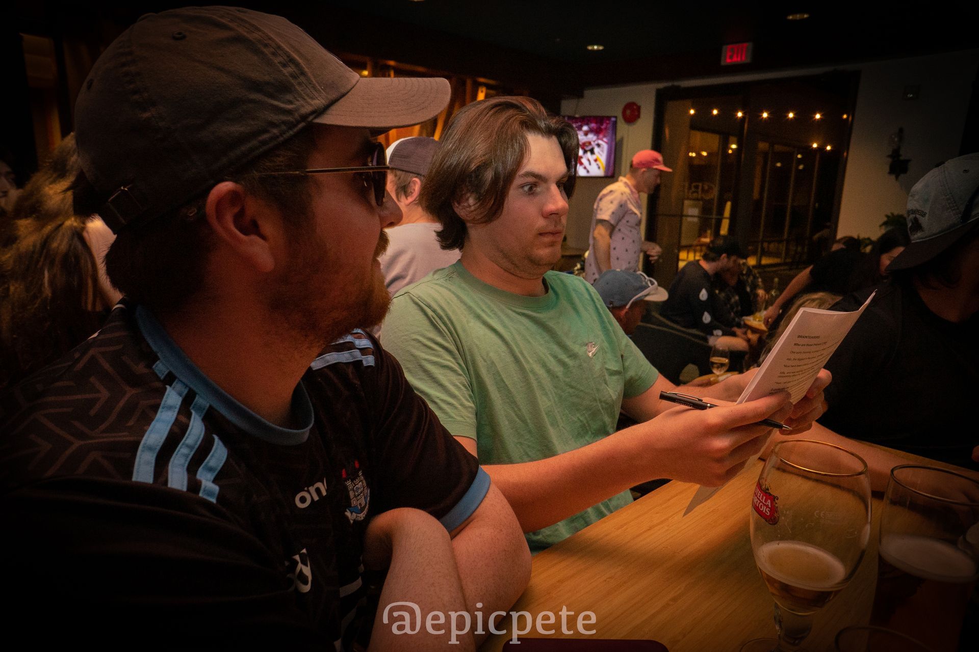 Two men are sitting at a table in a bar looking at a piece of paper.