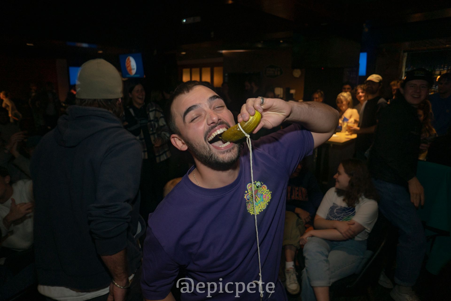 A man in a purple shirt is eating a pickle in a crowded room.