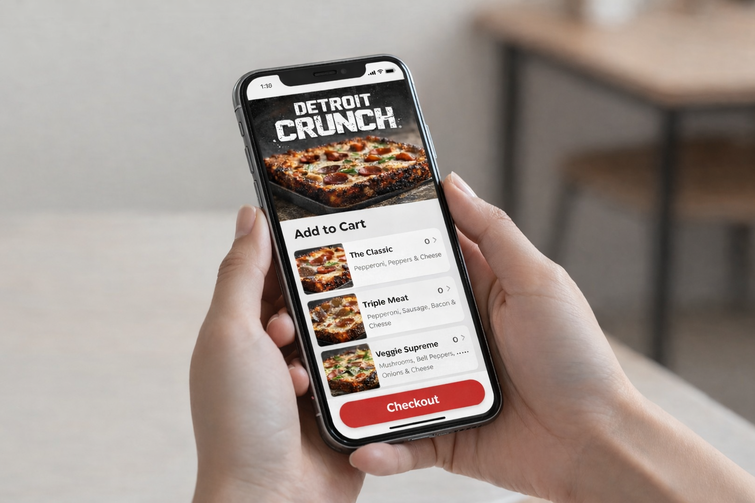 Hands holding a phone showing a Detroit Crunch pizza ordering app with pizza choices.