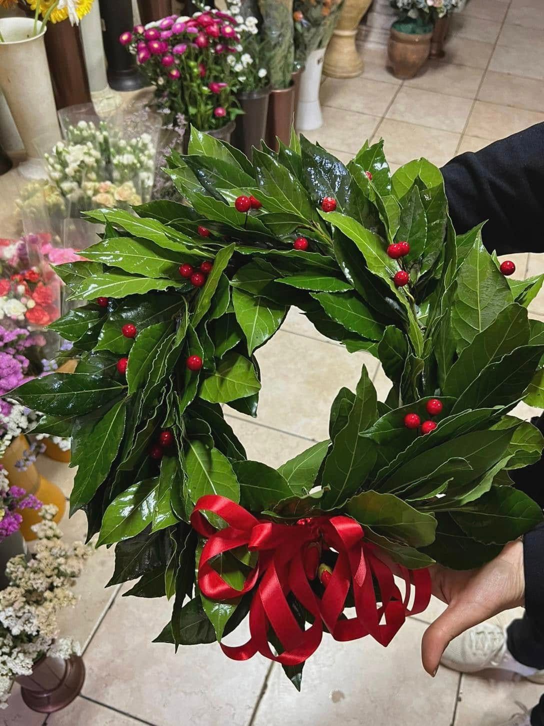 Corona d'alloro con bacche rosse e un nastro rosso, tenuta tra le mani da una persona, con fiori sullo sfondo.