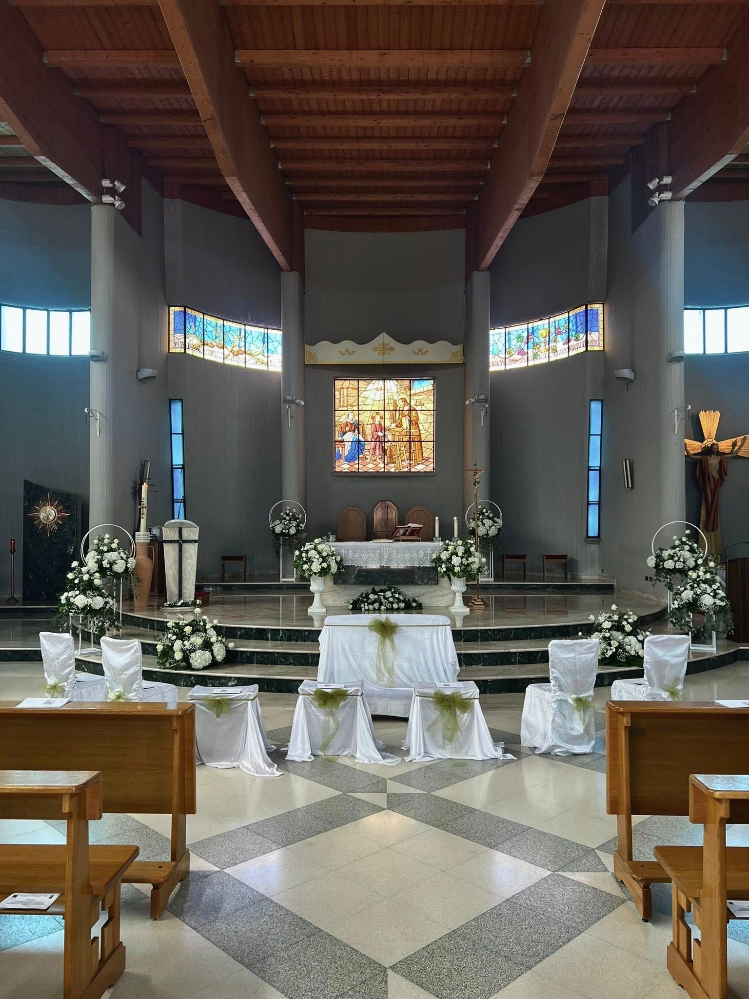 Interno della chiesa allestito per una cerimonia. Composizioni floreali bianche, tavolo dell'altare e sedie sono disposti in uno spazio ampio e luminoso.
