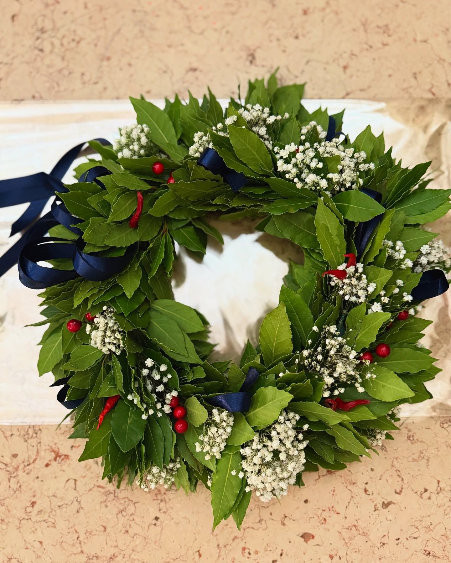Corona d'alloro con verde, piccoli fiori bianchi, bacche rosse e nastri blu navy.