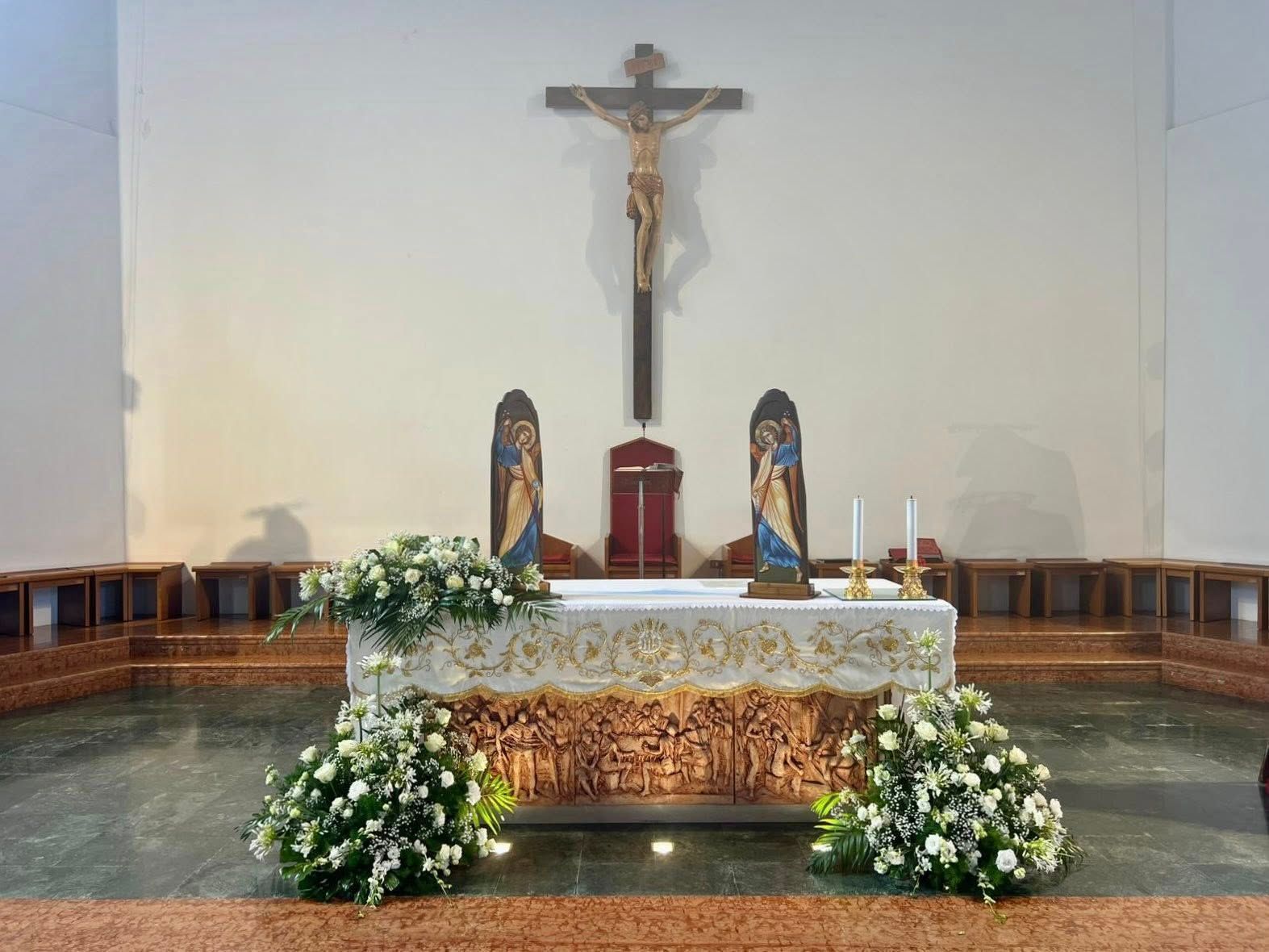 Altare in una chiesa con un crocifisso, composizioni floreali, statue di Maria e candele.