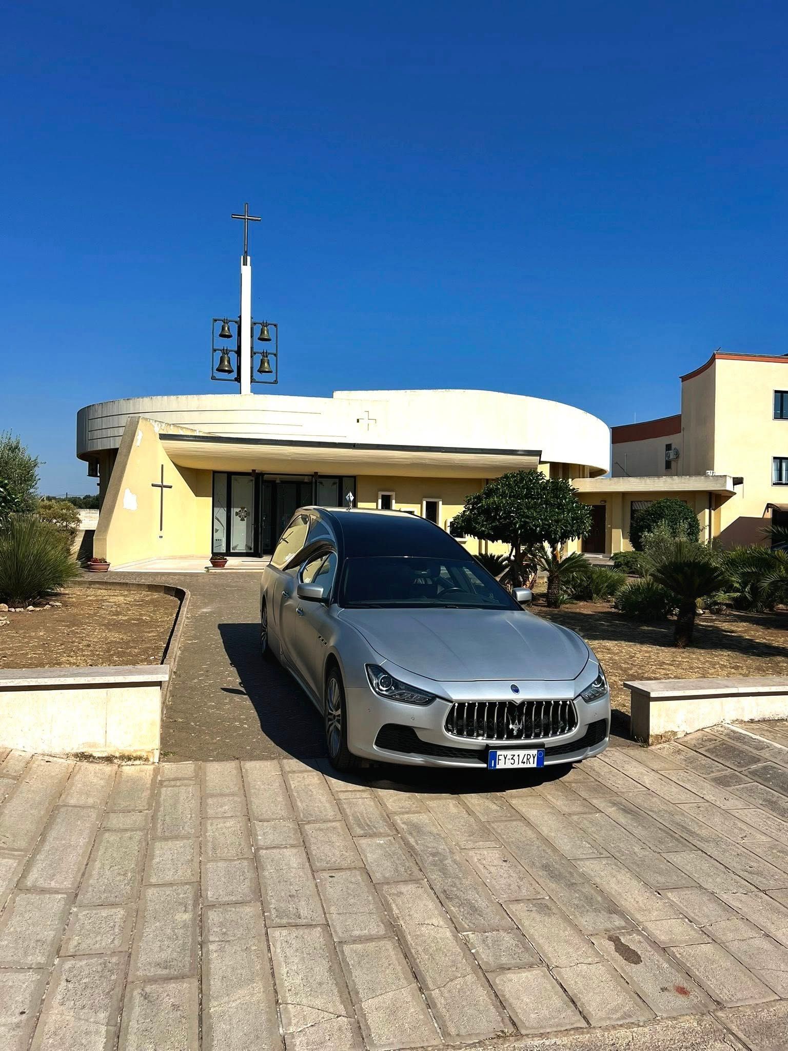 Una Maserati argentata parcheggiata davanti a un edificio chiaro con una croce, sotto un cielo azzurro e terso.