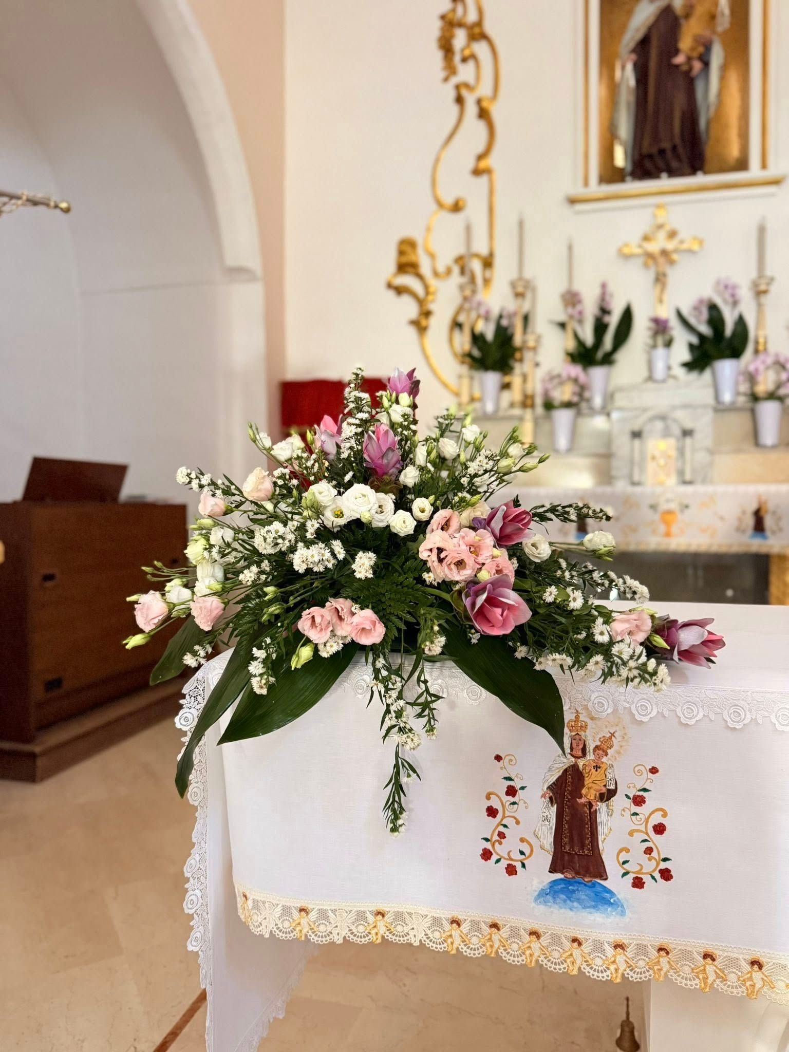 Composizione floreale su un altare in una chiesa. Fiori rosa e bianchi, dettagli dorati, sfondo religioso.