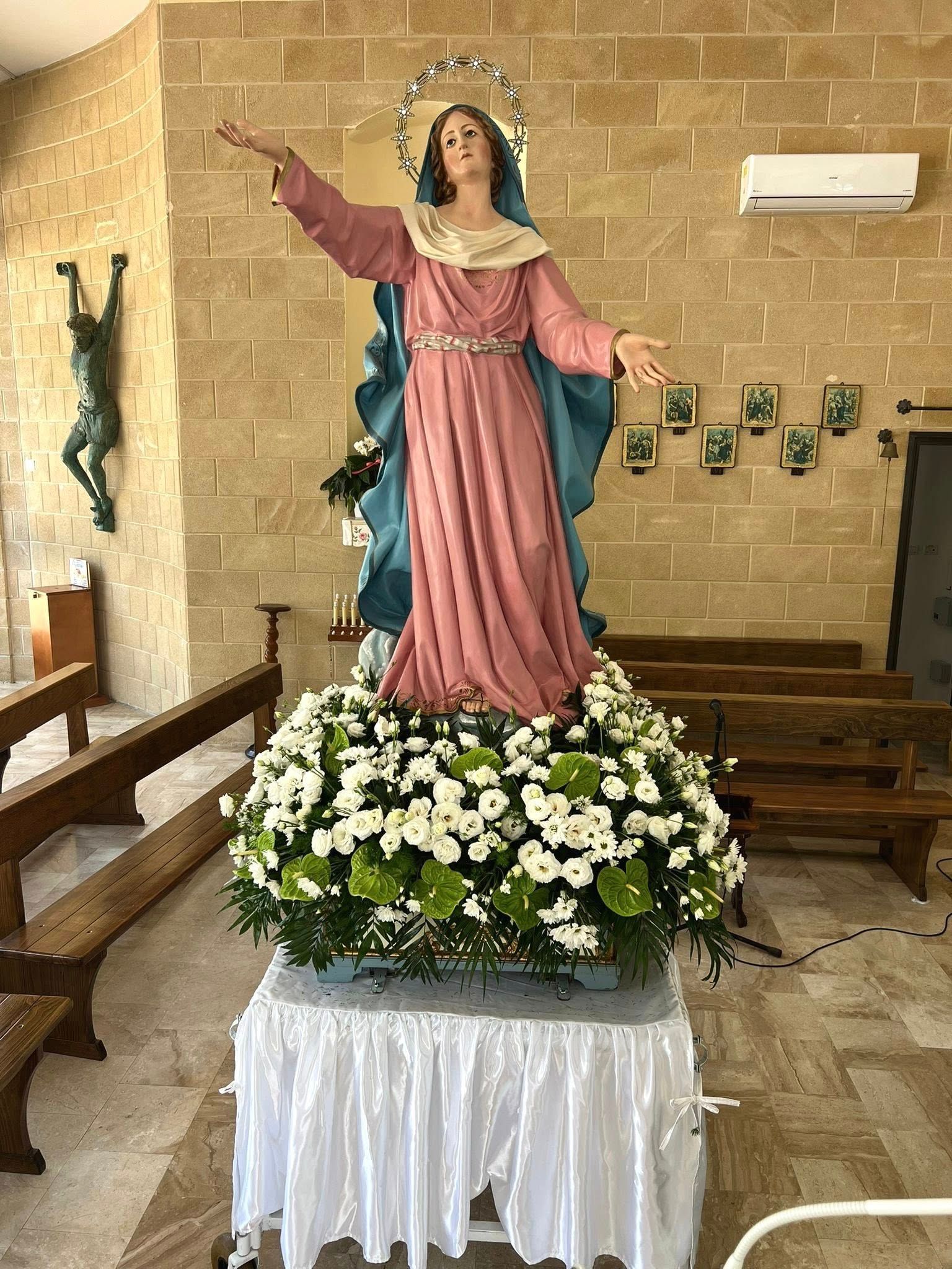 Statua della Vergine Maria, avvolta in un drappo rosa e blu, in cima a una composizione floreale, all'interno di una chiesa.