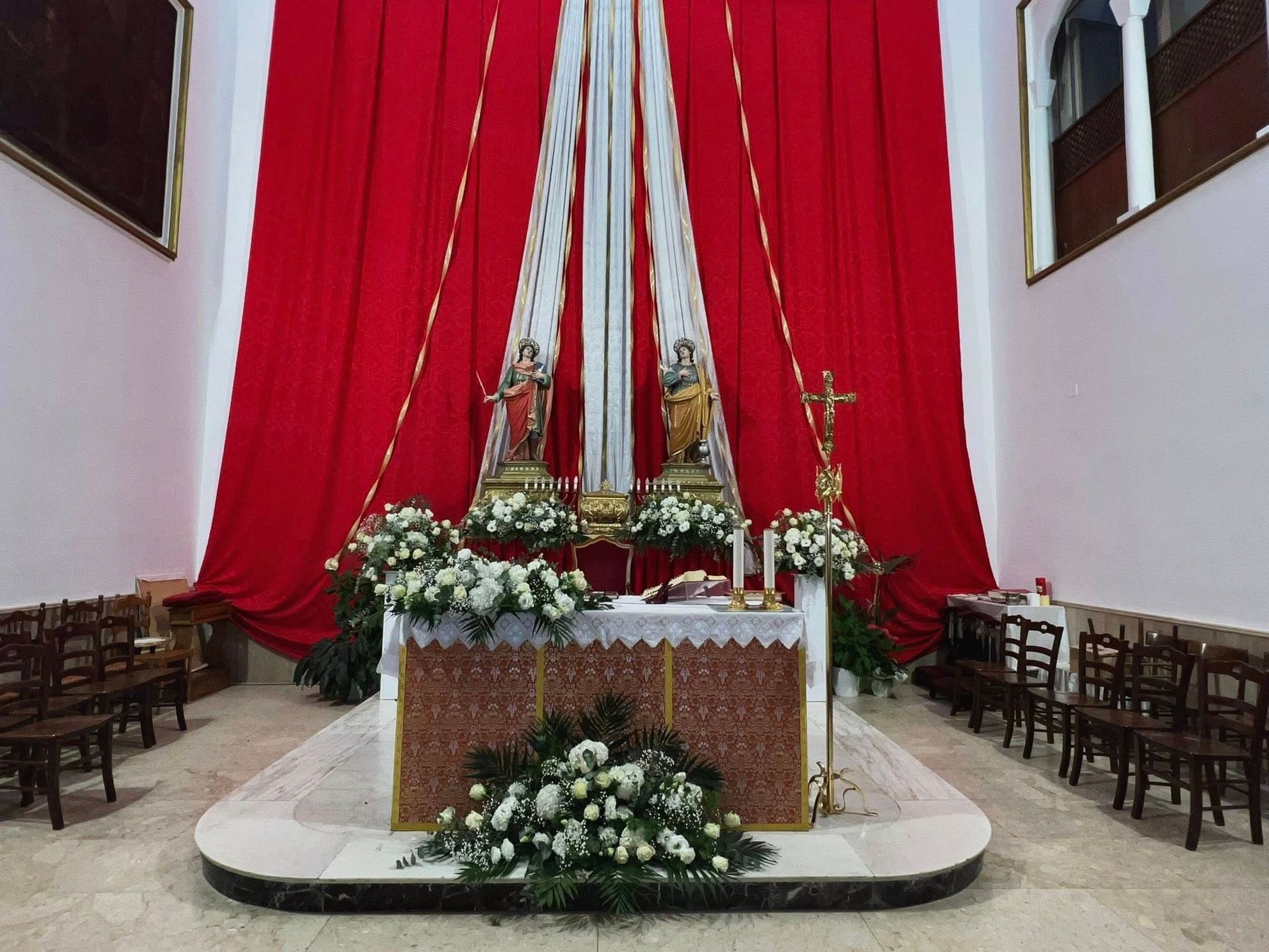 Altare della chiesa decorato con drappi rossi, fiori bianchi e statue, con file di sedie di legno.
