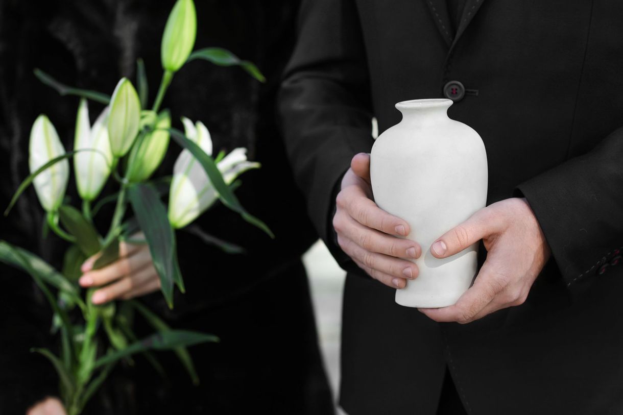 Persona in abito nero che tiene in mano un'urna bianca, accanto a una persona che tiene in mano dei gigli bianchi.