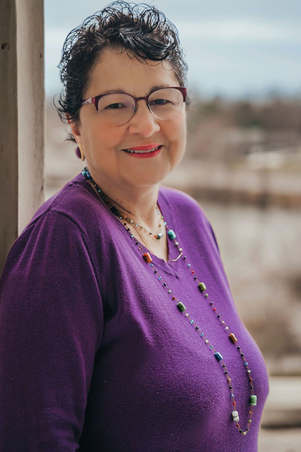 A woman wearing a purple sweater and glasses is smiling for the camera.
