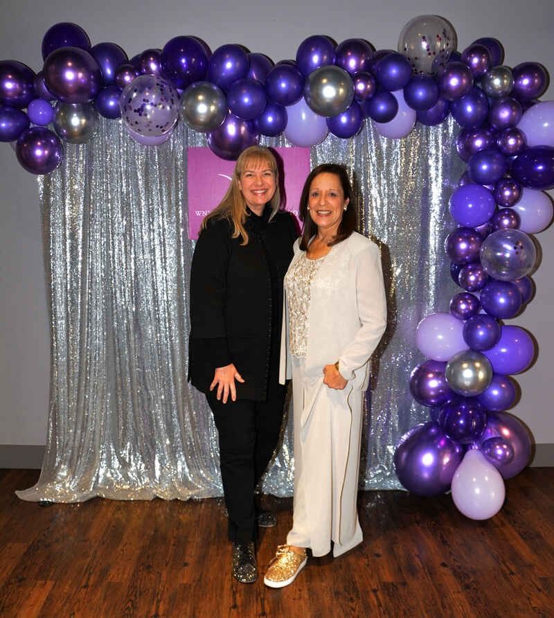 Two women are standing in front of a purple balloon arch.