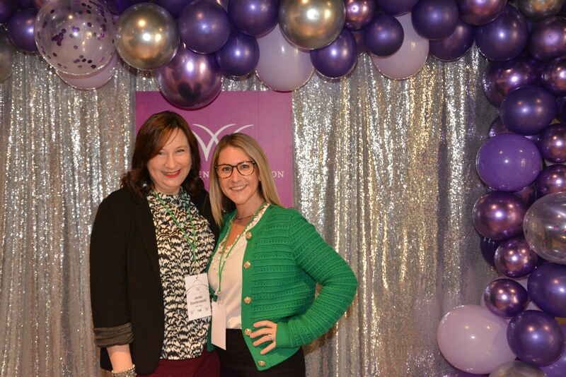 Two women are posing for a picture in front of a wall of purple balloons.