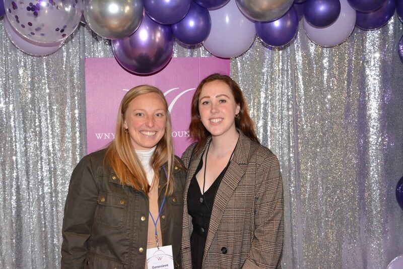 Two women are posing for a picture in front of purple balloons.