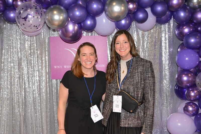 Two women are posing for a picture in front of purple balloons.