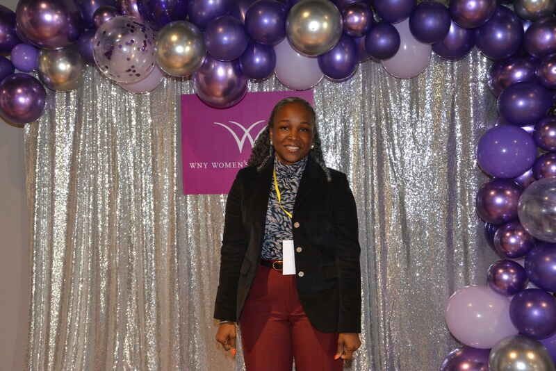 A woman is standing in front of a wall of purple and gold balloons.