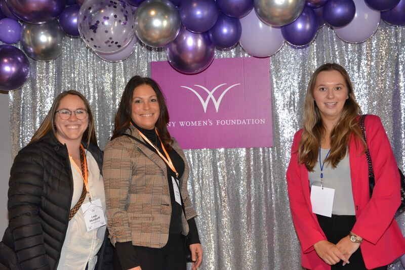 Three women are posing for a picture in front of purple and silver balloons.