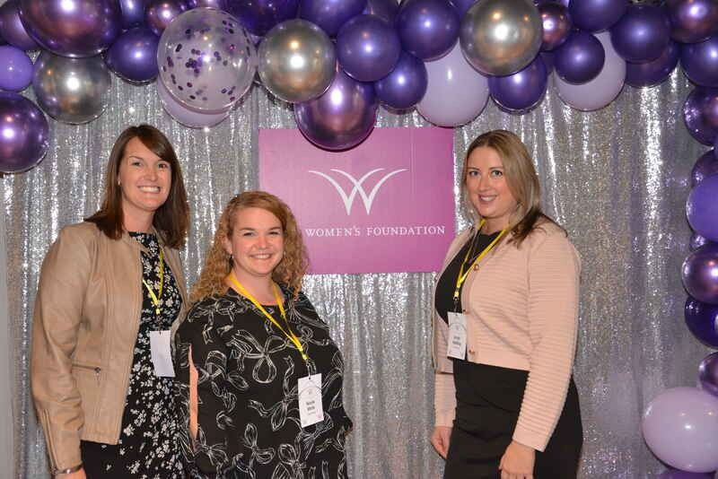 Three women are posing for a picture in front of a wall of purple balloons.