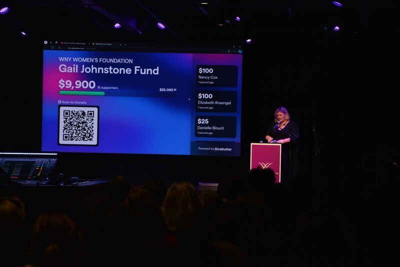 A woman is giving a speech at a podium in front of a large screen.