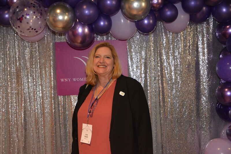 A woman is standing in front of a purple and gold balloon arch.