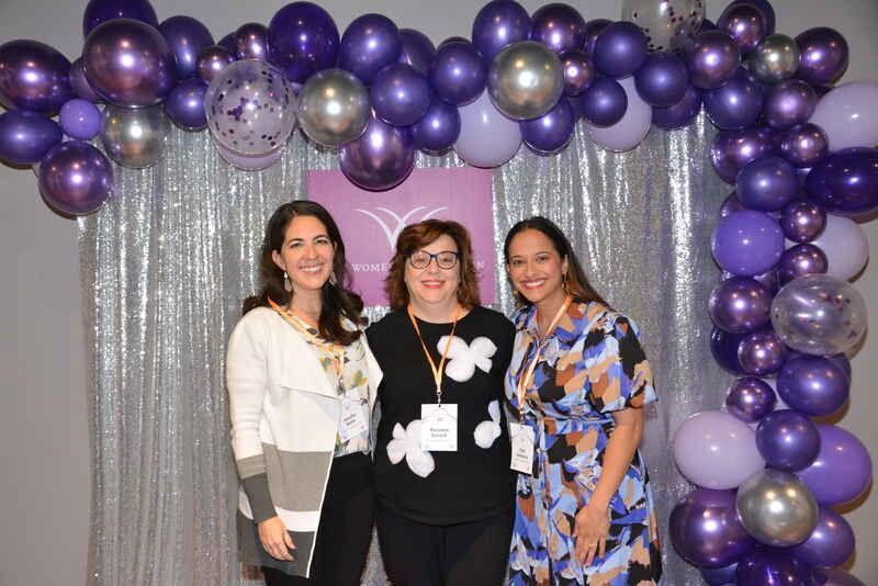 Three women are posing for a picture in front of a purple balloon arch.