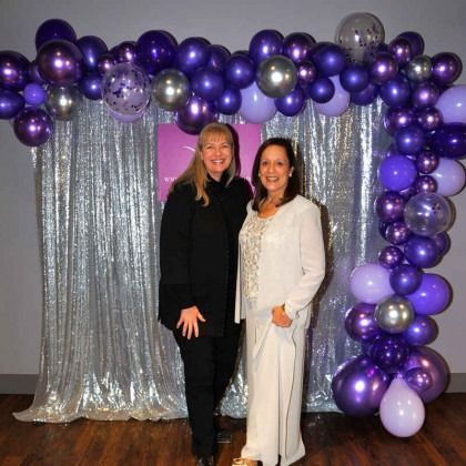 Two women are standing in front of a purple balloon arch