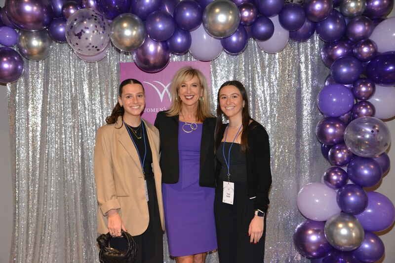 Three women are posing for a picture in front of a purple balloon arch.