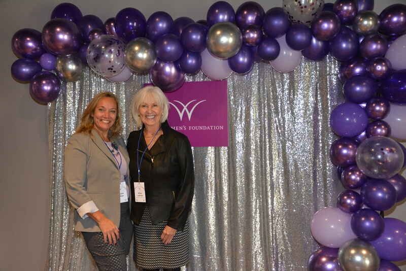 Two women are posing for a picture in front of a purple balloon arch.